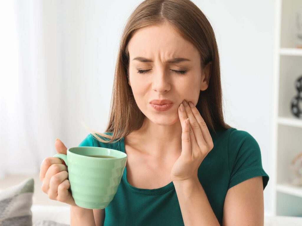 Woman in green shirt wincing in pain, holding her cheek and a green mug of coffee, suggesting a toothache or discomfort while drinking.