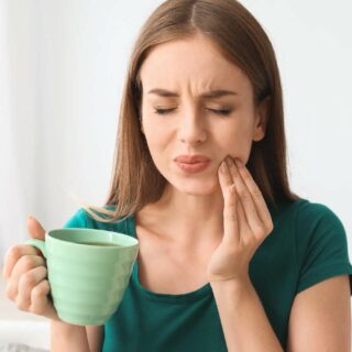 Woman in green shirt wincing in pain, holding her cheek and a green mug of coffee, suggesting a toothache or discomfort while drinking.