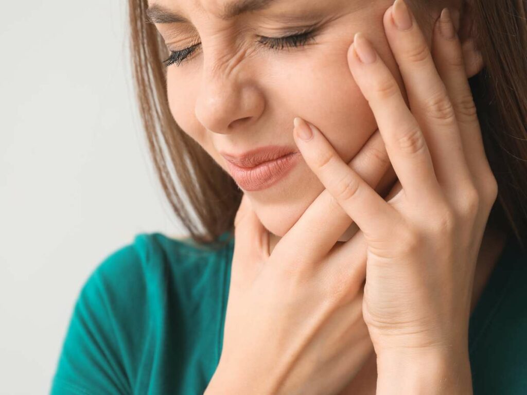 A woman in a green top touches her cheek and temple, appearing to be in tooth pain or discomfort.