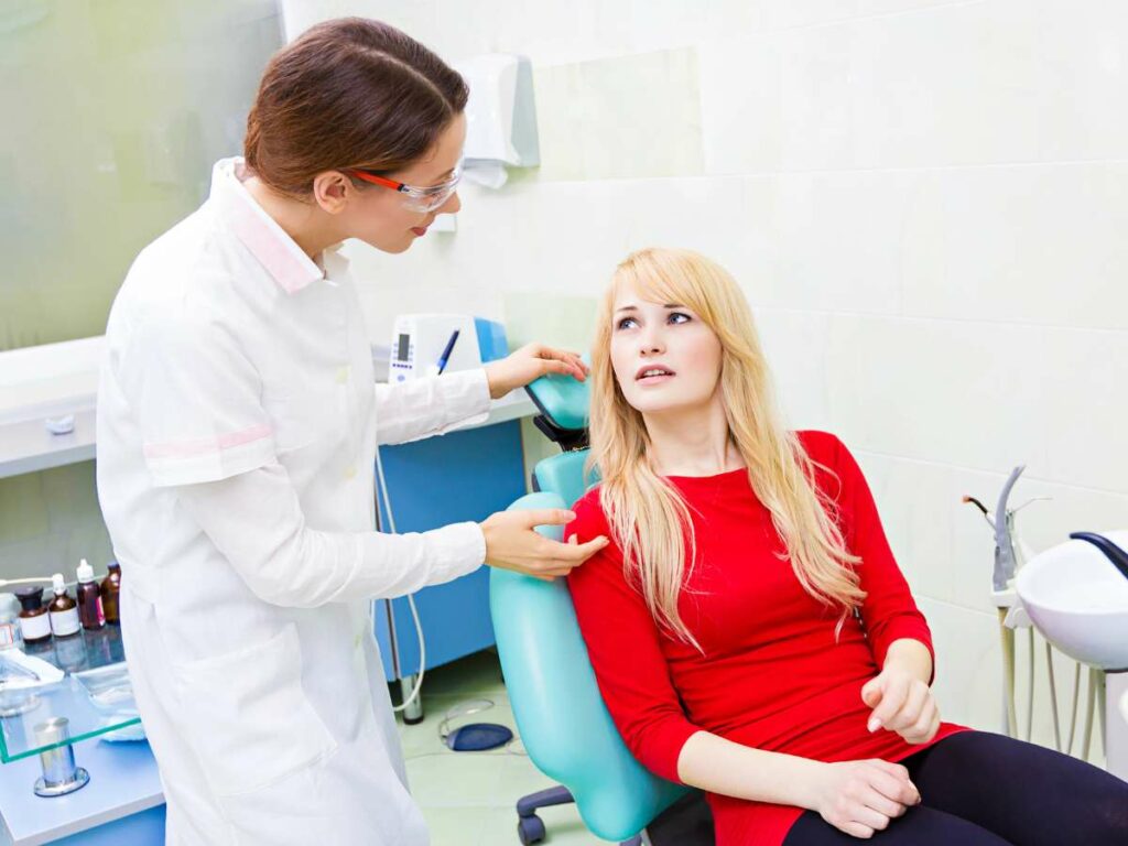 Dentist in a white coat comforting a worried female patient in a red shirt sitting in a dental chair in a clinic.