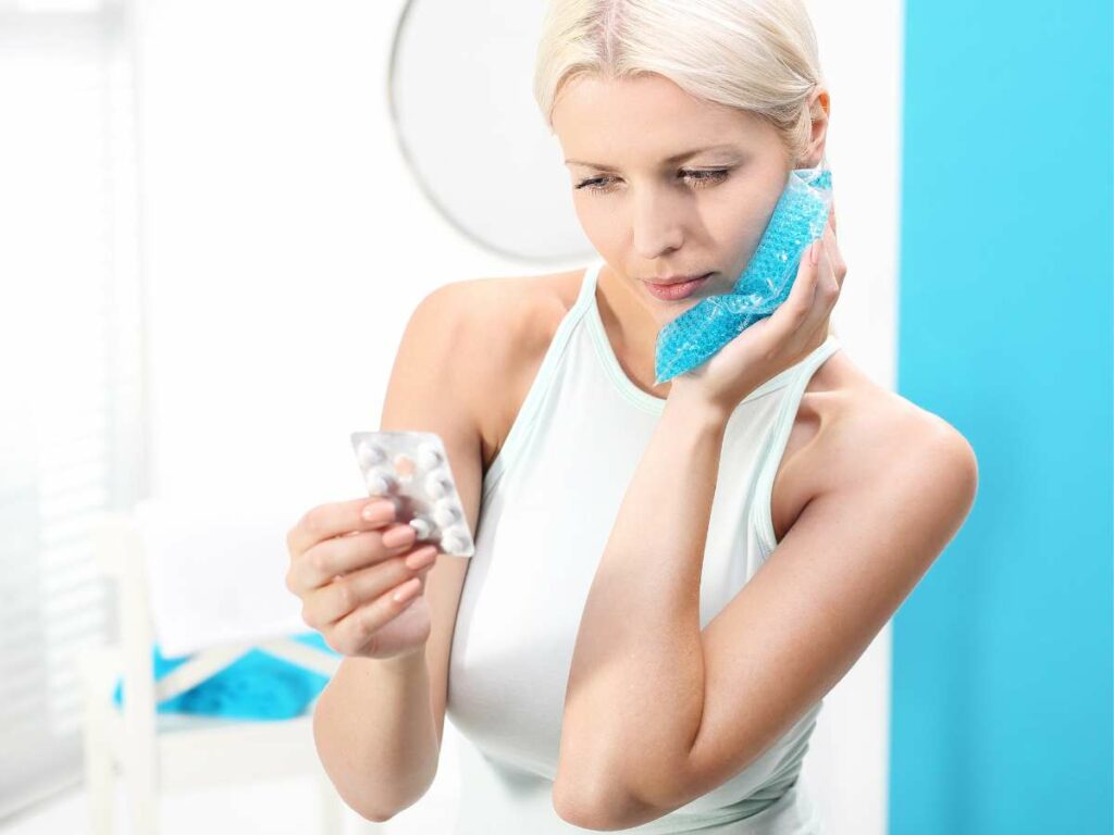 A woman holds a pack of pills while applying an ice pack to her cheek, appearing to manage pain or discomfort.