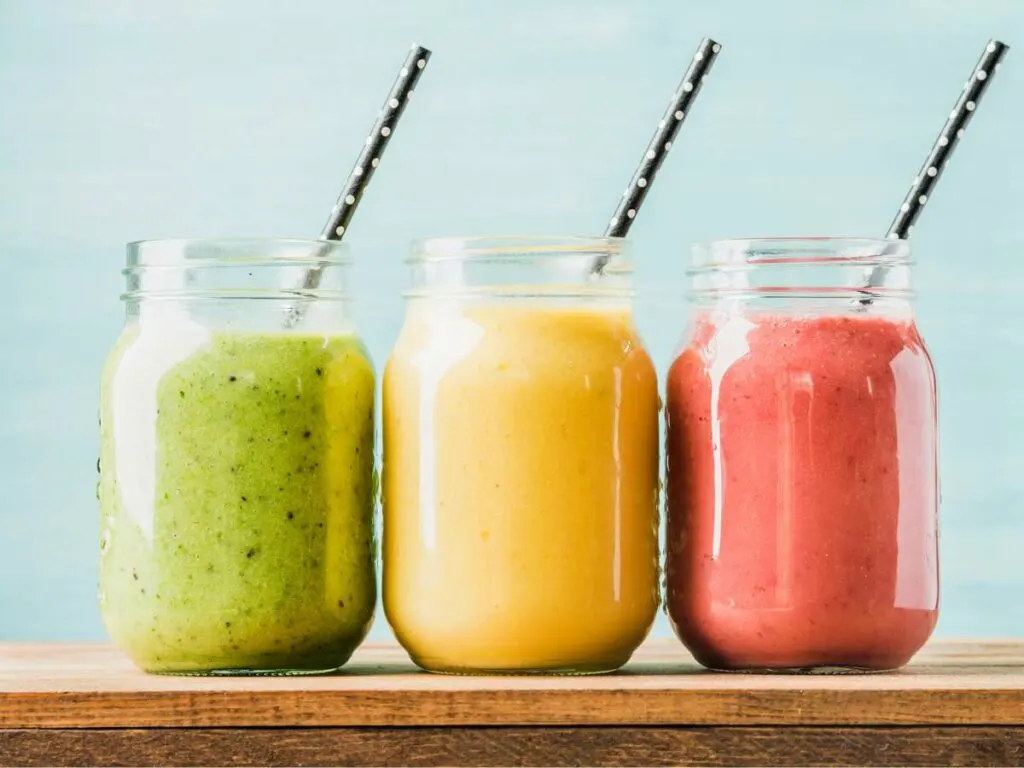 Three jars of smoothies with straws, in green, yellow, and red colors, aligned on a wooden table against a blue background.