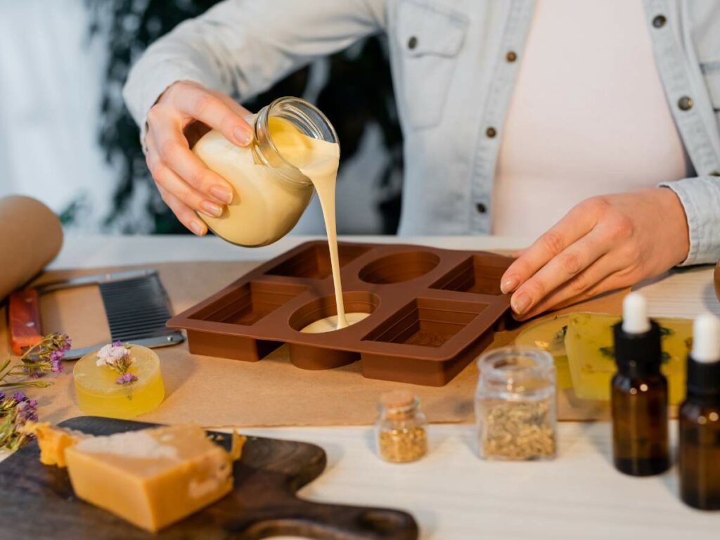 A person pours liquid soap base into a mold, surrounded by soap-making ingredients and tools on a table.