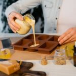 A person pours liquid soap base into a mold, surrounded by soap-making ingredients and tools on a table.