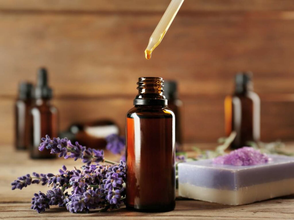 A dropper dispensing essential oils into a small amber bottle surrounded by lavender flowers and handmade soap on a wooden surface.