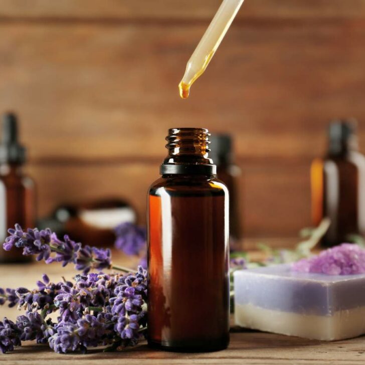 A dropper dispensing essential oils into a small amber bottle surrounded by lavender flowers and handmade soap on a wooden surface.