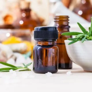A bottle of essential oil with fresh rosemary and other herbs in the background.