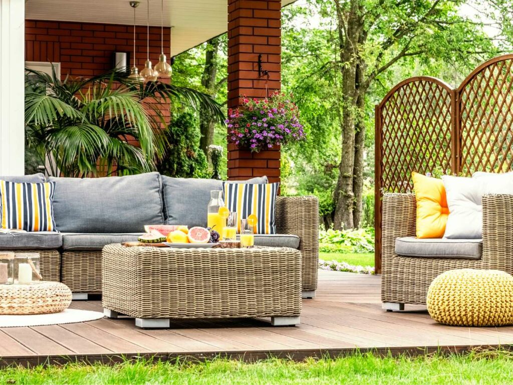 Outdoor patio setting for entertaining with rattan furniture, cushions, and a table with refreshments, surrounded by greenery and a brick house.