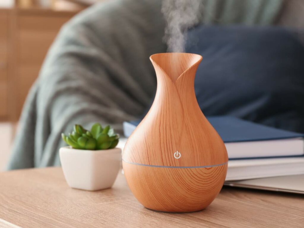 A wooden essential oil diffuser releasing vapor on a table next to a small green plant and books, with a blurred background.