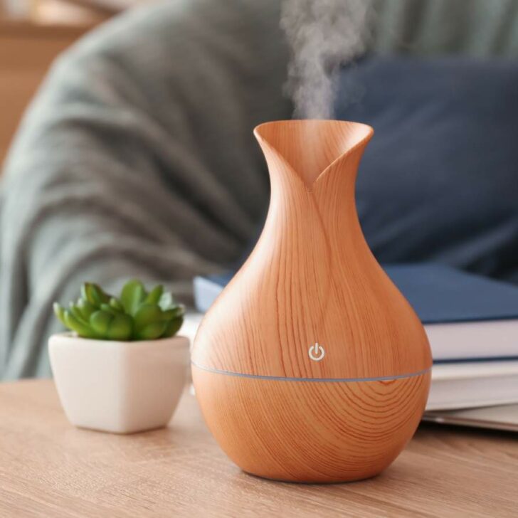 A wooden essential oil diffuser releasing vapor on a table next to a small green plant and books, with a blurred background.