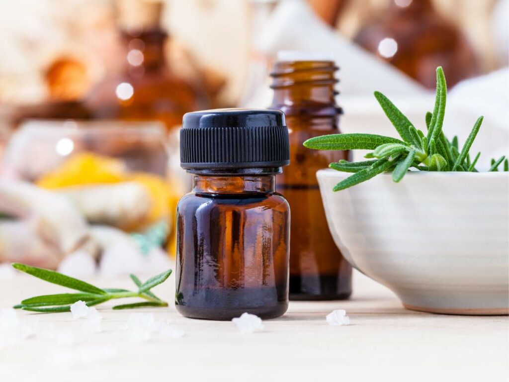 A small brown glass bottle with essential oil next to a bowl of fresh rosemary and scattered salt crystals on a wooden surface.