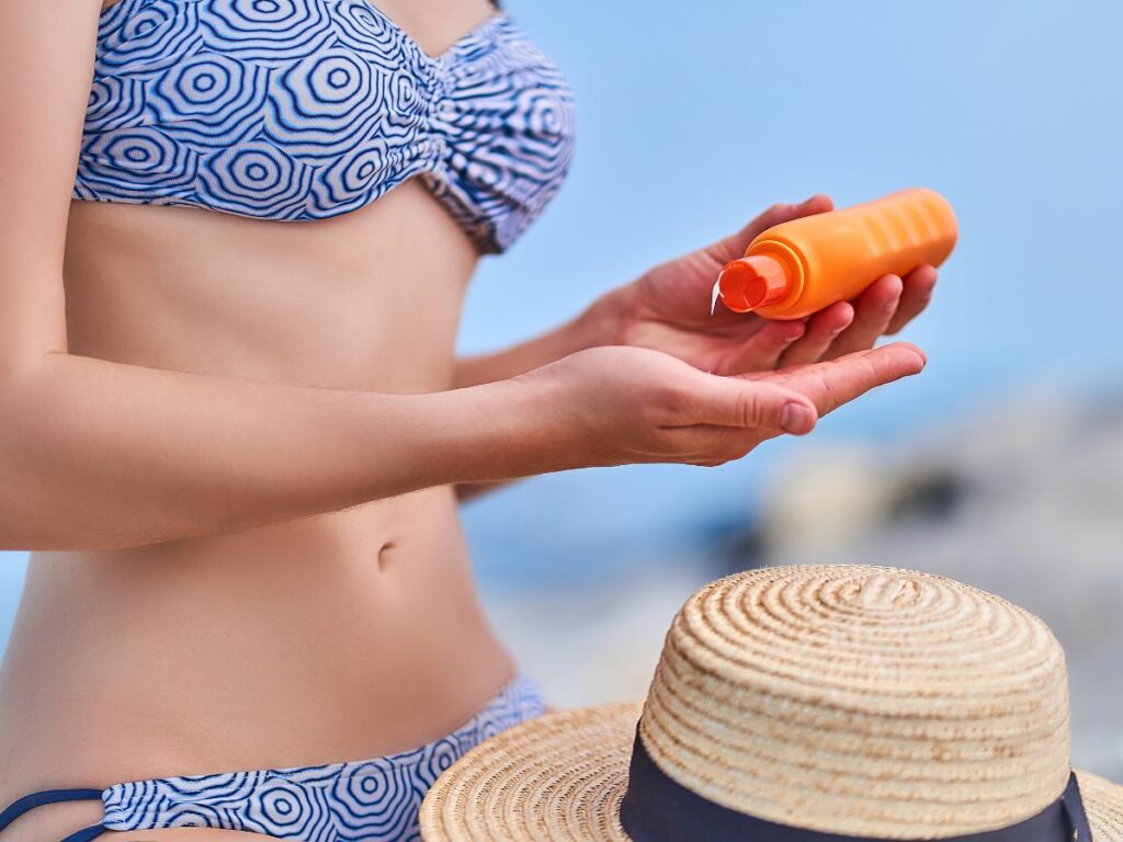A woman wearing a bikini applies sunscreen lotion from an orange bottle on a sunny beach, with a straw hat nearby.