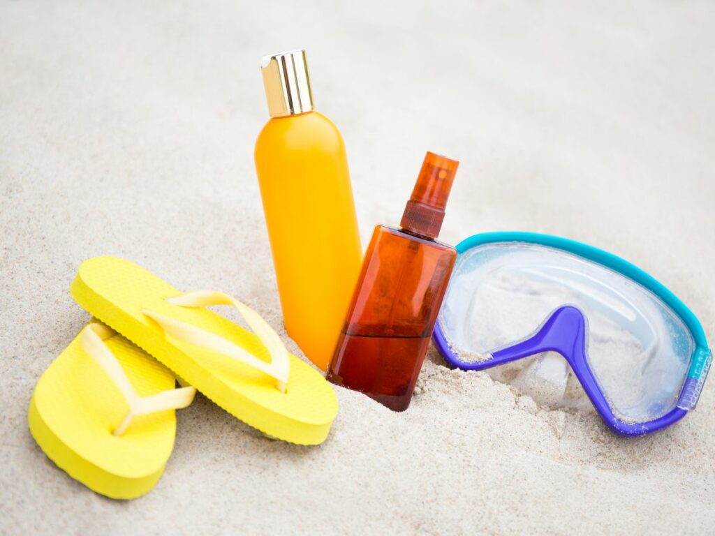 A pair of yellow flip-flops, two suntan lotion bottles, and blue snorkeling goggles on a sandy beach.
