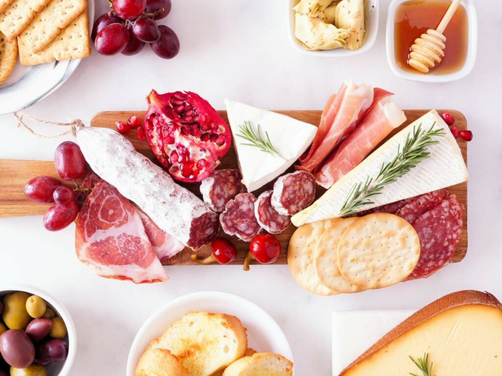 A charcuterie board with an assortment of meats, cheeses, crackers, and fruit, accompanied by small bowls of olives and honey, arranged on a white surface.