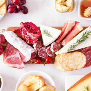 A charcuterie board with an assortment of meats, cheeses, crackers, and fruit, accompanied by small bowls of olives and honey, arranged on a white surface.