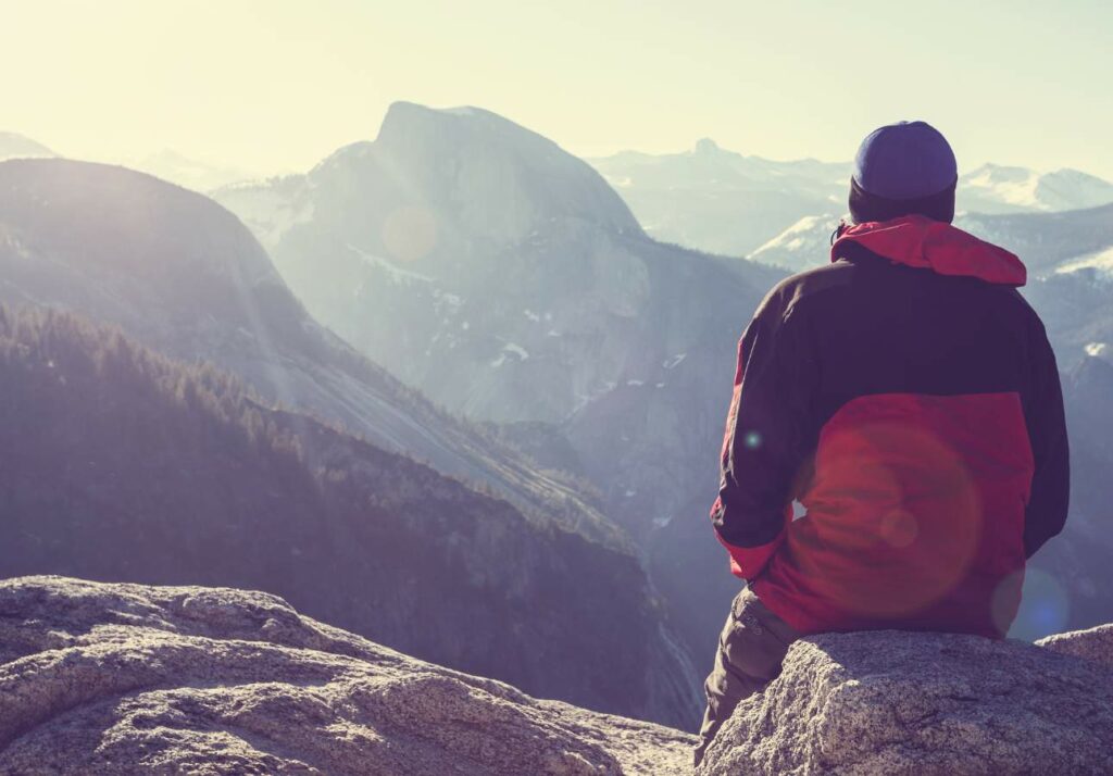 Person in a red and black jacket sitting on a rock overlooking a mountainous landscape with sun rays and distant peaks.