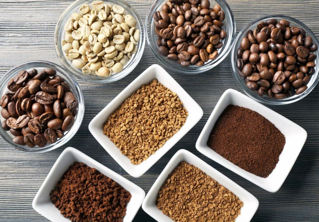 Assortment of coffee products in bowls on a wooden surface, including whole beans, ground coffee, and instant coffee granules.