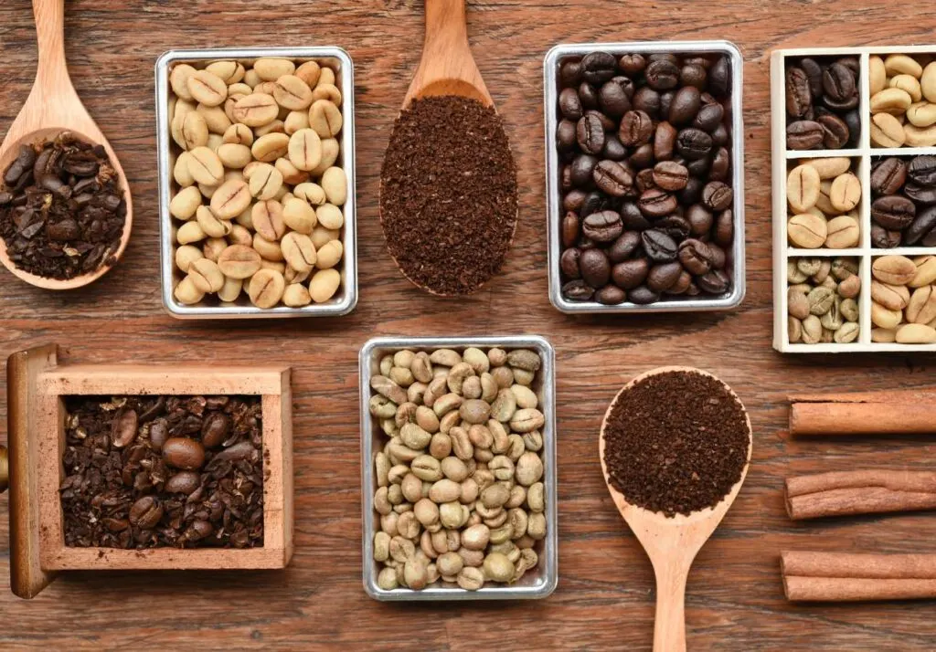 Various types of coffee beans and ground coffee displayed in boxes and spoons on a wooden surface, accompanied by cinnamon sticks.
