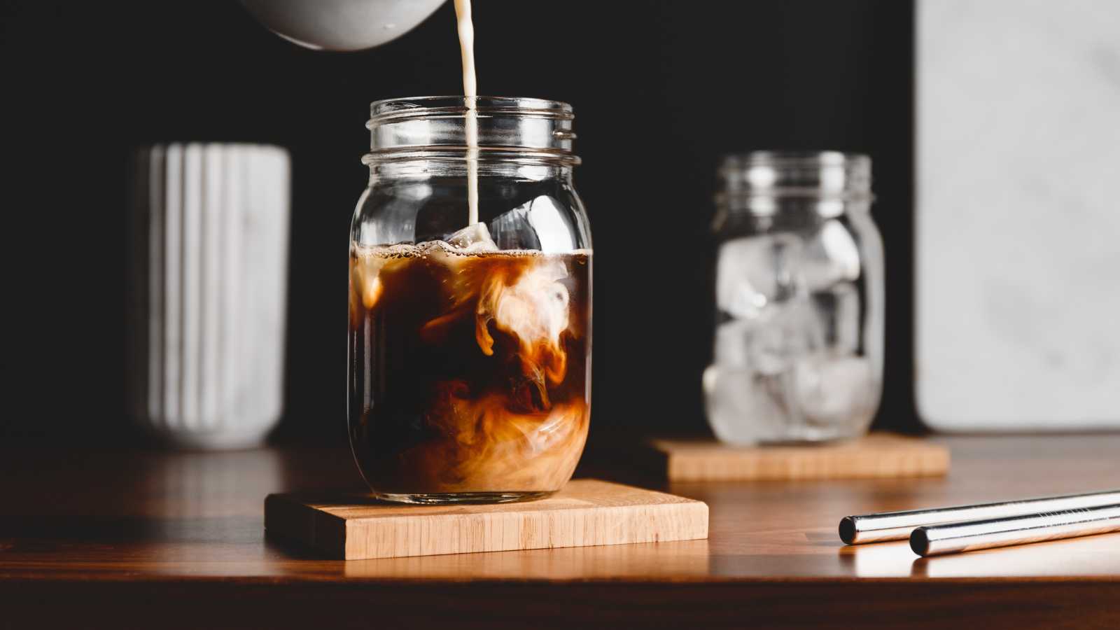 Milk being poured into a glass jar of cold brew coffee on a wooden coaster.