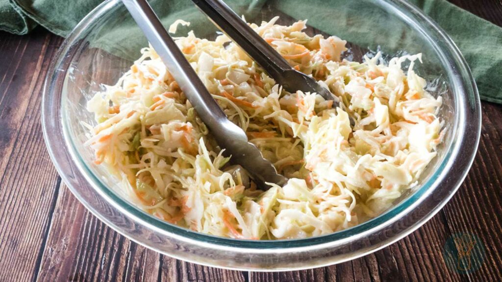 A glass bowl filled with coleslaw, containing shredded cabbage and carrots, with metal tongs resting on top.