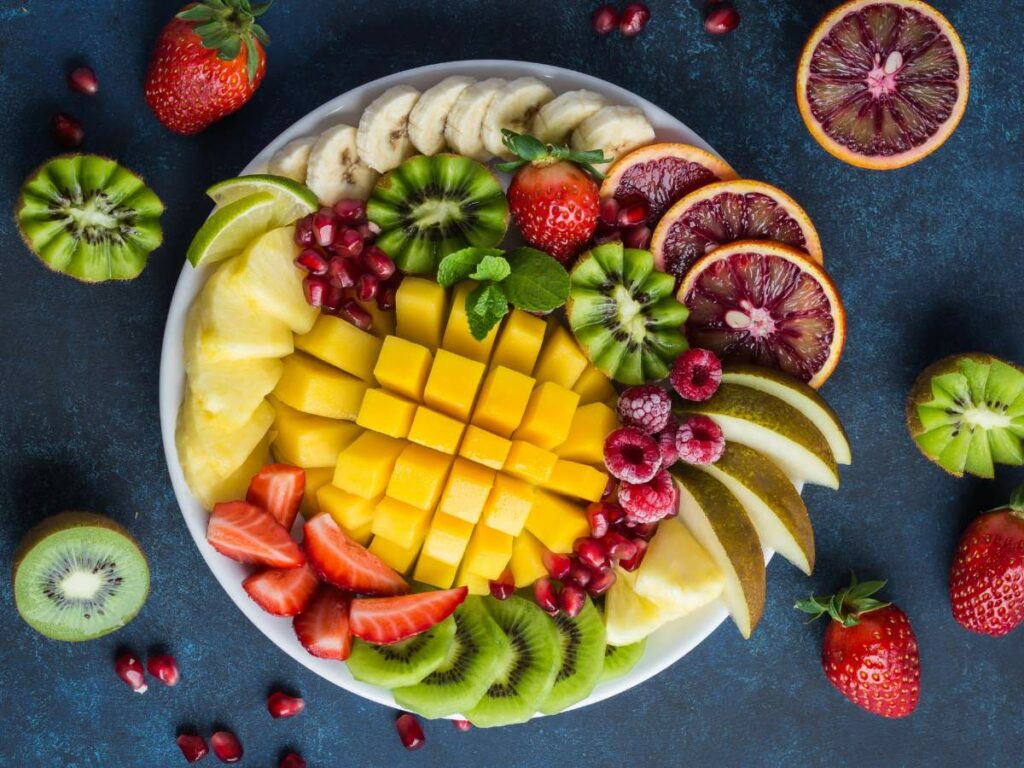 A plate arranged with various sliced fruits including mango, strawberries, pomegranate, banana, kiwi, pineapple, pear, lime, and blood orange on a dark blue surface.