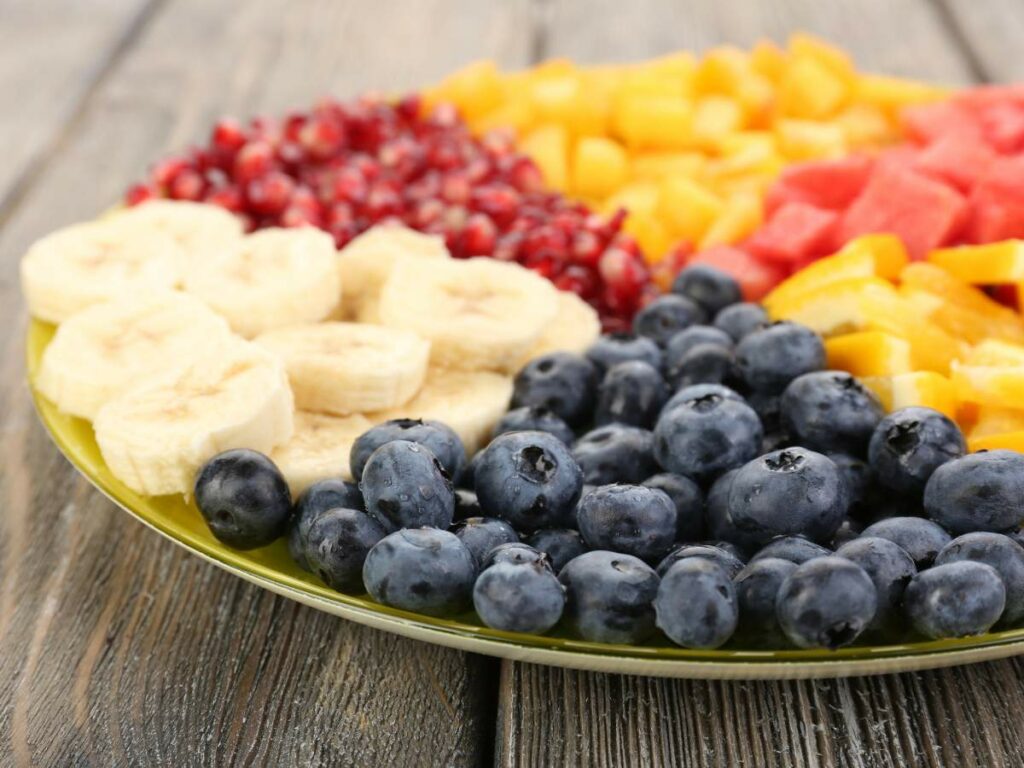 A plate of assorted fresh fruits including blueberries, banana slices, pomegranate seeds, pineapple chunks, and watermelon pieces on a wooden surface.