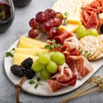 A small charcuterie board with slices of cheese, cured meats, grapes, blackberries, crackers, and herbs.