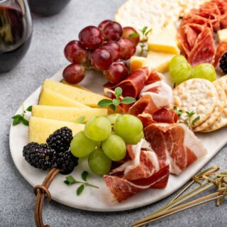 A small charcuterie board with slices of cheese, cured meats, grapes, blackberries, crackers, and herbs.