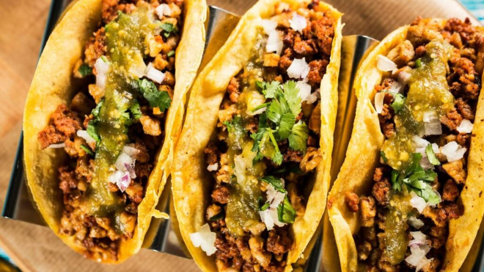 Three tacos filled with seasoned ground meat, chopped onions, cilantro, and topped with green salsa, arranged on a metal taco holder.