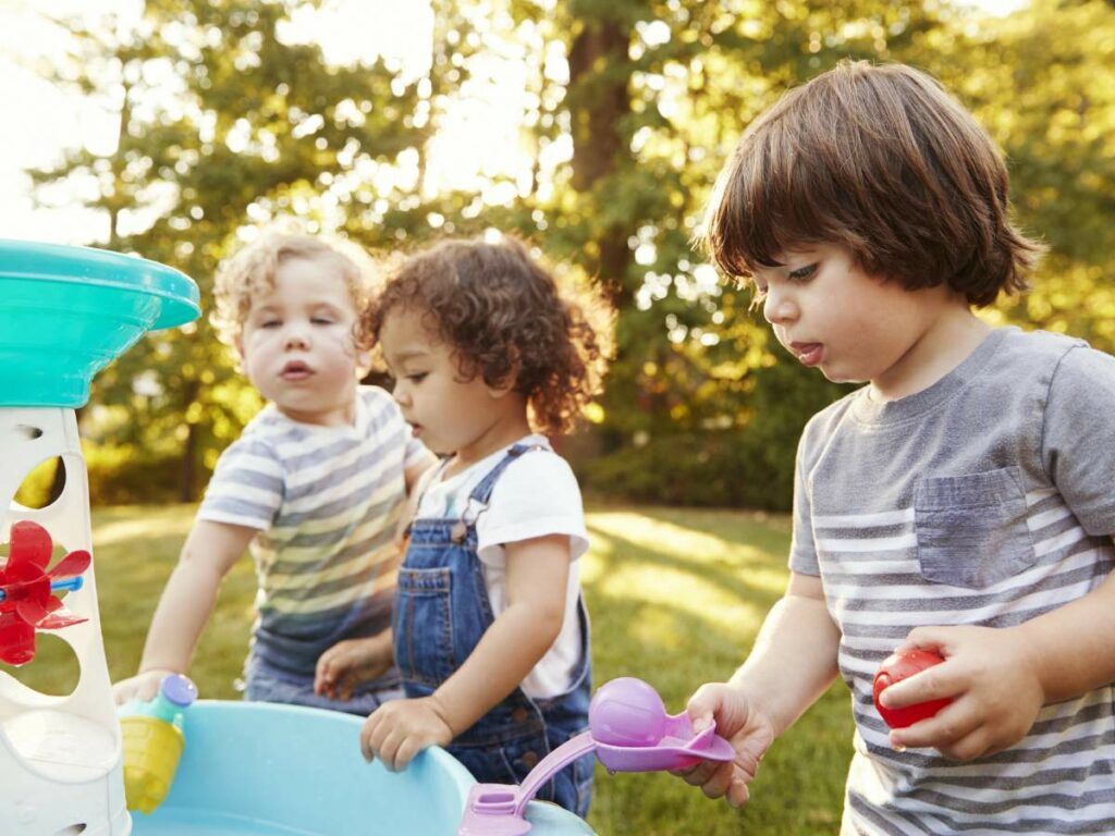 Three young children play outdoors with a water table, surrounded by greenery.