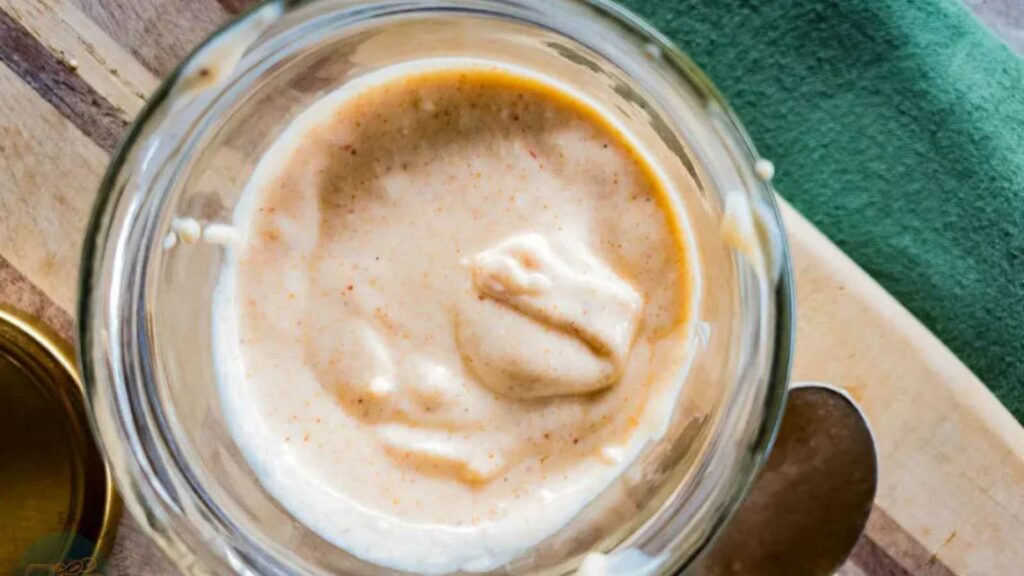 A close-up view of tiger sauce in a glass jar placed on a wooden surface next to a green cloth and a metal spoon.