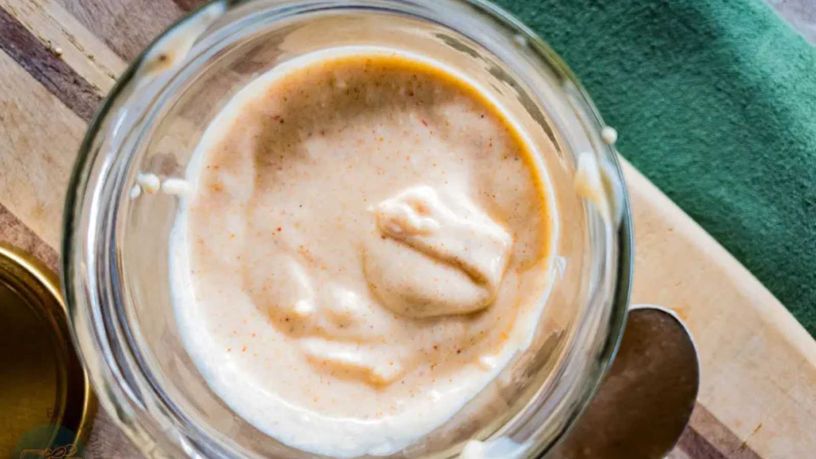 A close-up view of tiger sauce in a glass jar placed on a wooden surface next to a green cloth and a metal spoon.