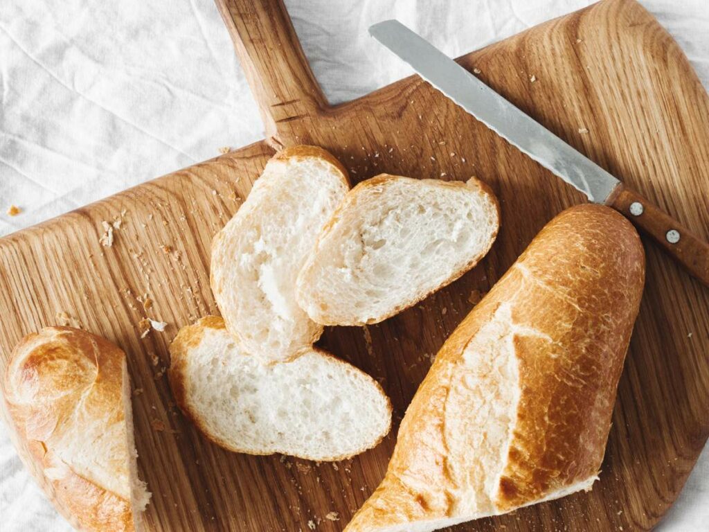 A wooden cutting board with a partially sliced baguette and a knife.