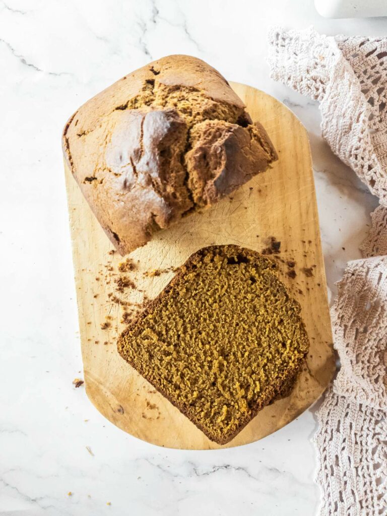 A loaf of homemade pumpkin bread sits on a wooden cutting board with one slice cut.