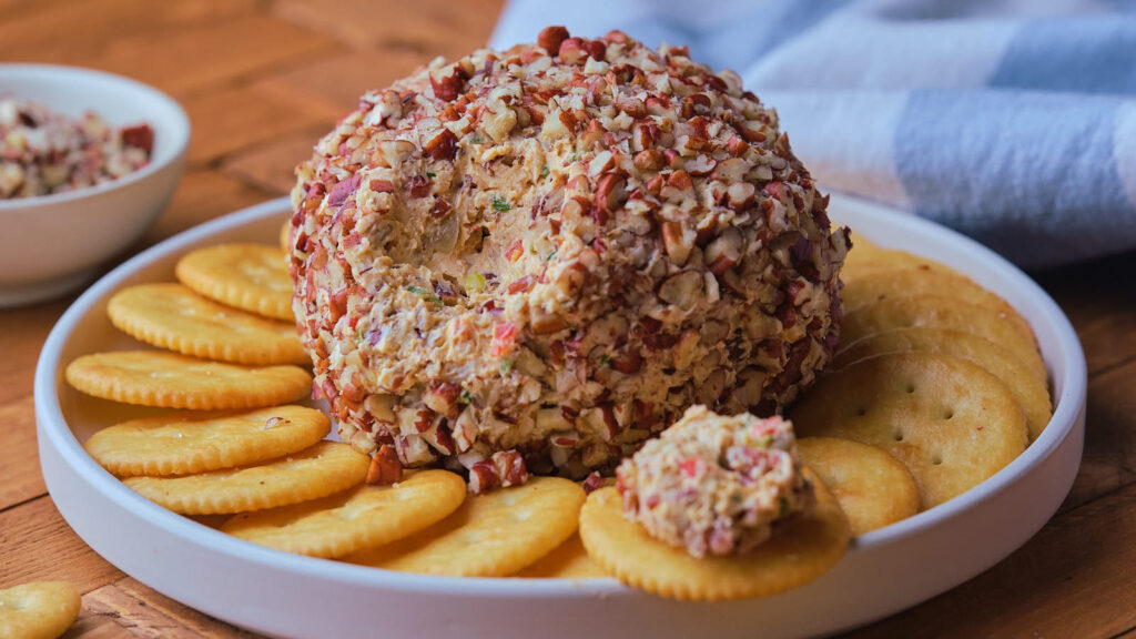 Pineapple cheese ball coated with chopped nuts is placed on a white plate surrounded by round crackers.