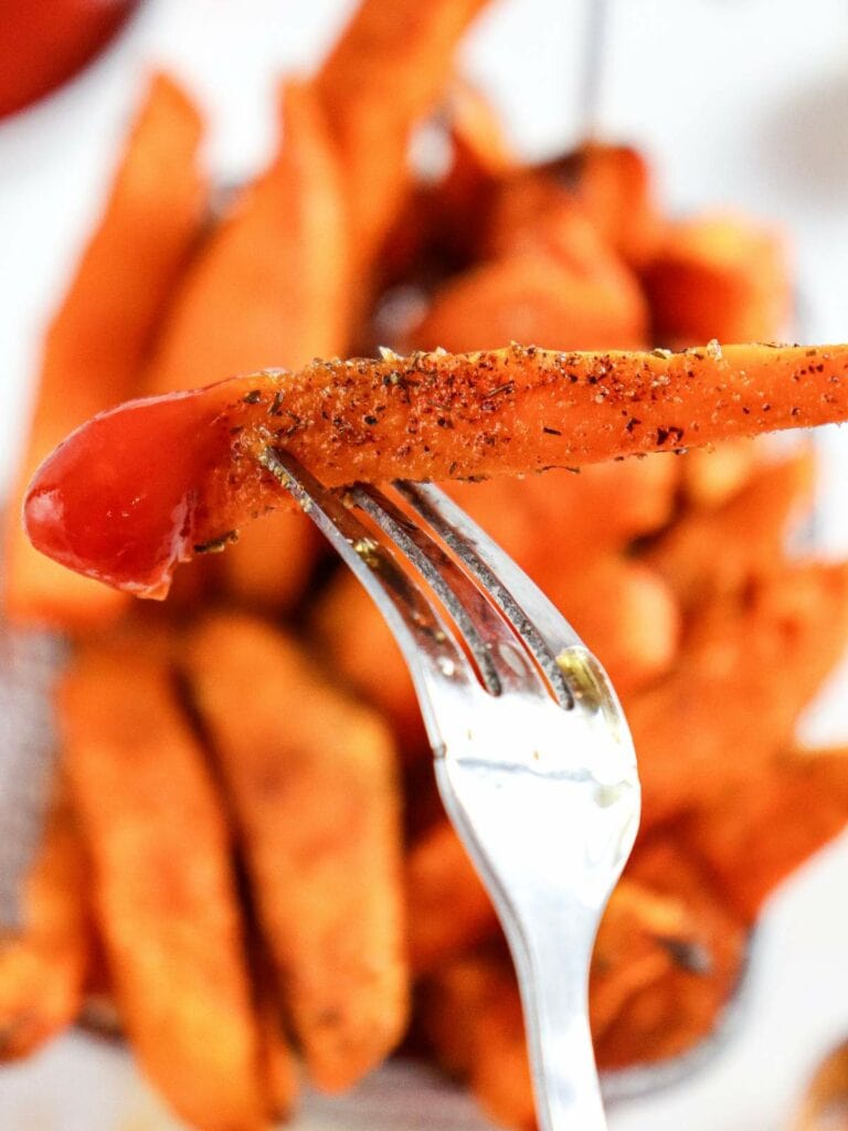 A fork holding a seasoned sweet potato fry dipped in ketchup, with more sweet potato fries in the background.