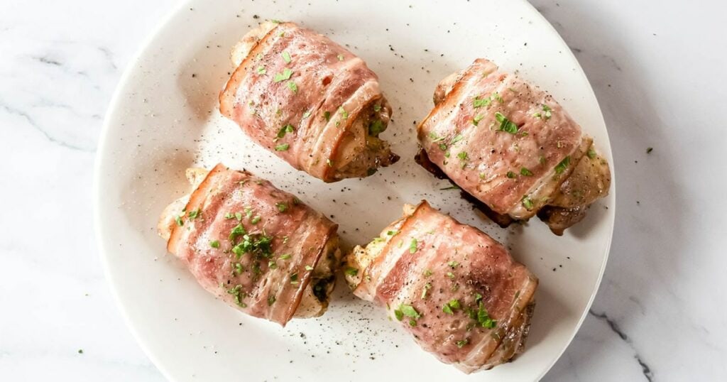 Four bacon-wrapped chicken pieces garnished with chopped herbs on a white plate.