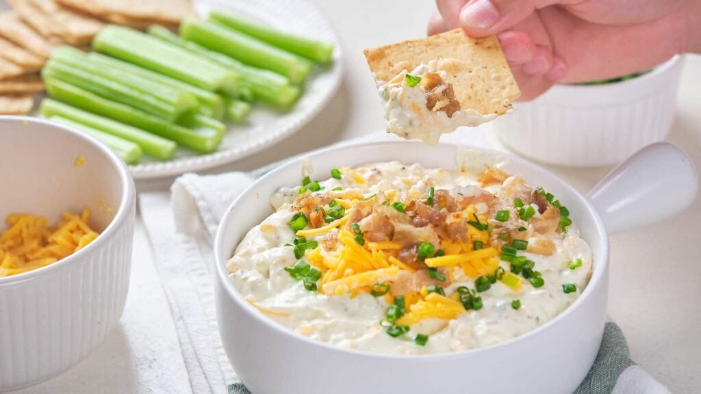 A hand dips a cracker into a bowl of crack dip topped with bacon, cheese, and chives.