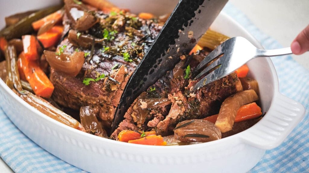 Slow cooker pot roast in a white baking dish. A fork and knife are being used to slice the meat.