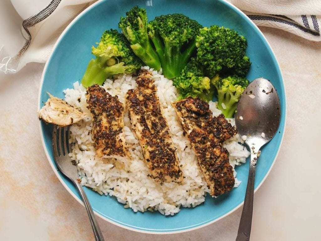 A blue plate with sliced seasoned chicken, white rice, and broccoli. A fork and spoon are placed beside the food.