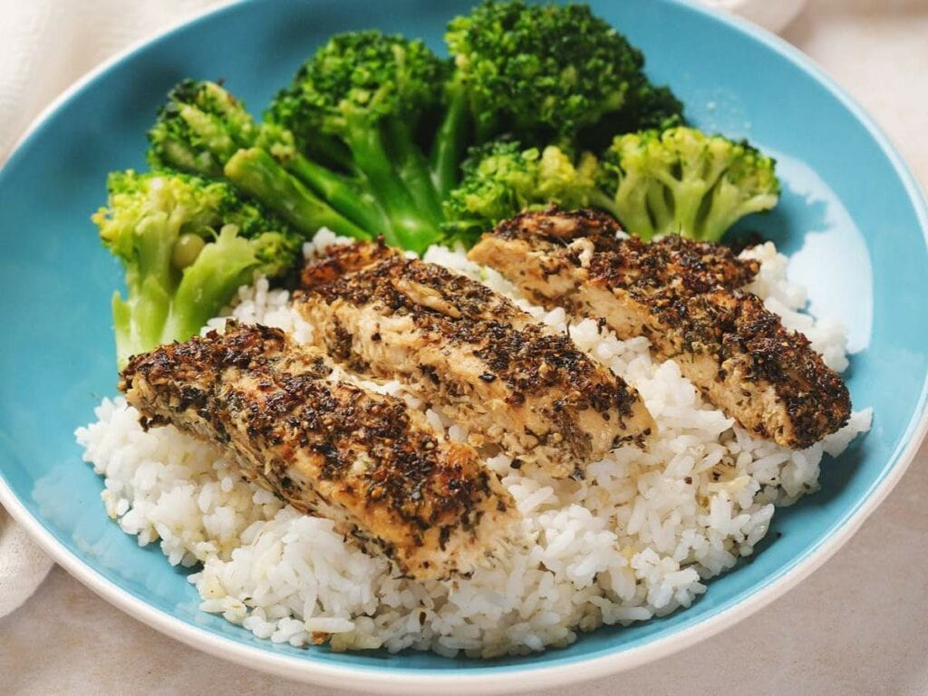 A plate with herb-crusted chicken strips on white rice, served with steamed broccoli.