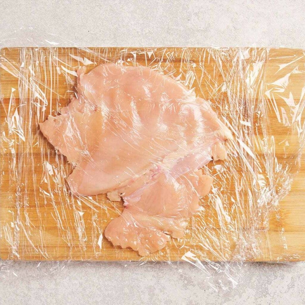 Flattened raw chicken breast under plastic wrap on a wooden cutting board.