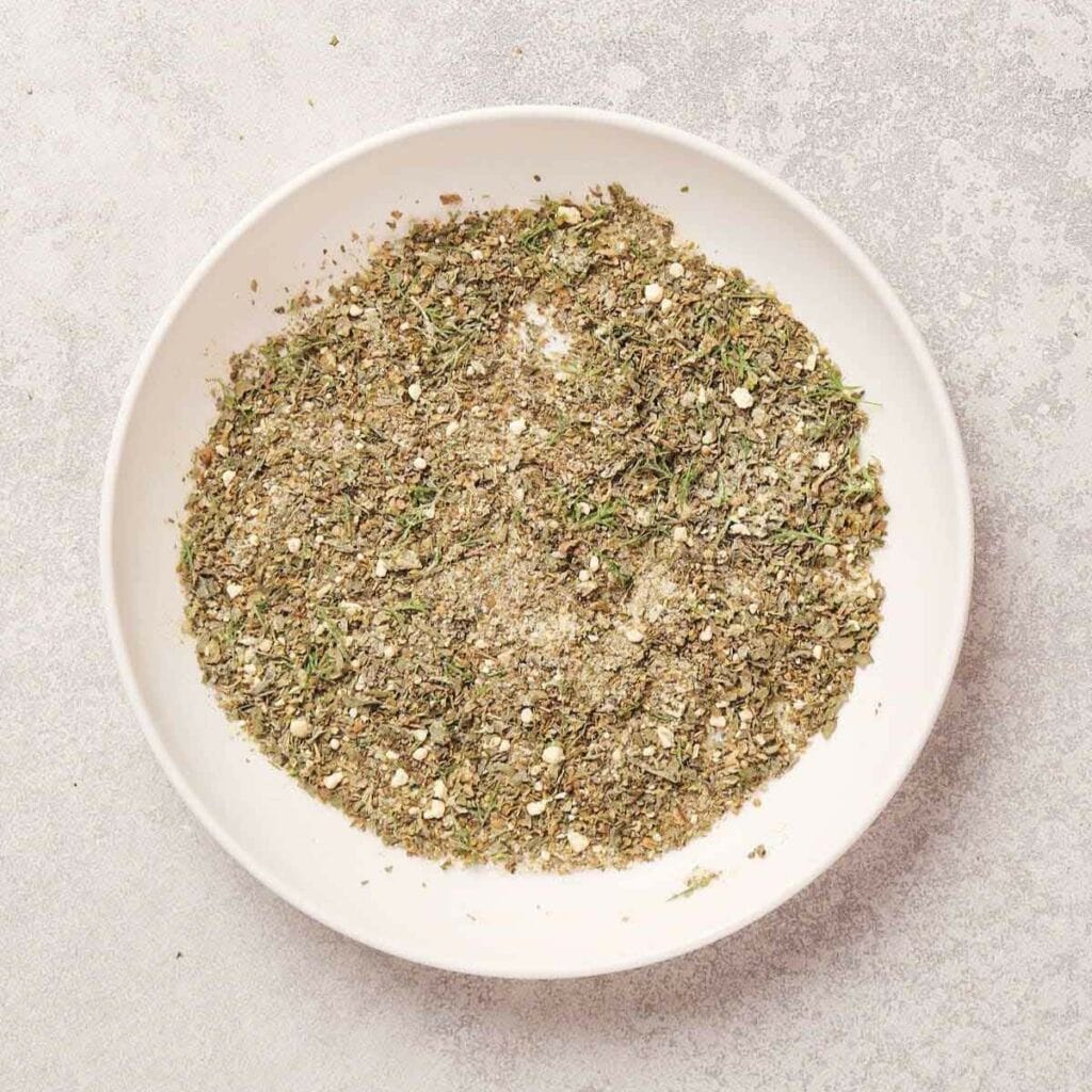 A white bowl containing a mixed blend of dried herbs and spices on a light gray surface.