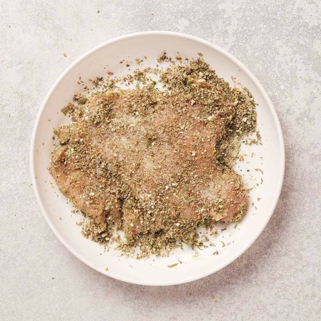 Raw chicken breast coated with mixed herbs and spices on a white plate.