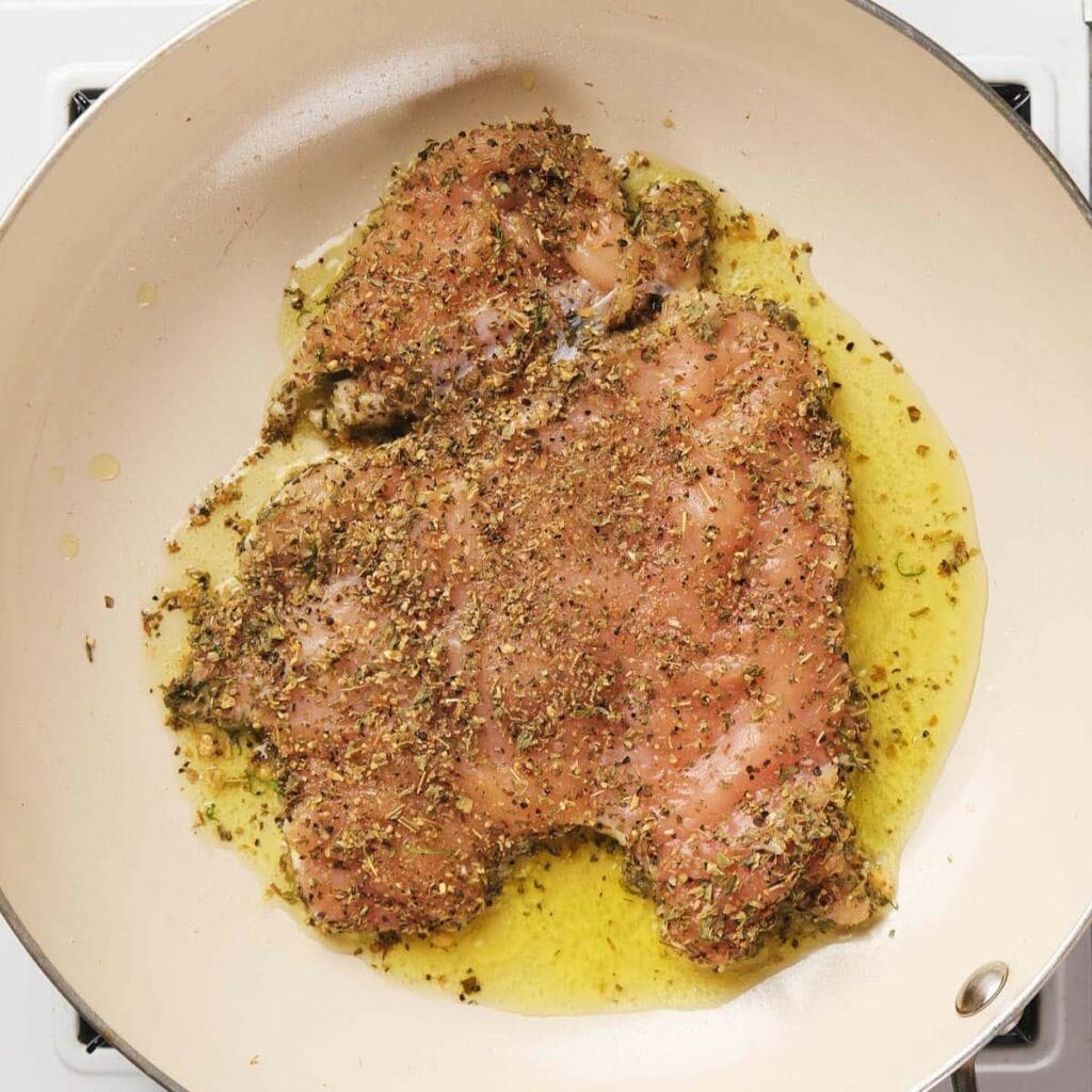 Chicken breast seasoned with herbs and spices, cooking in olive oil in a frying pan on a stovetop.