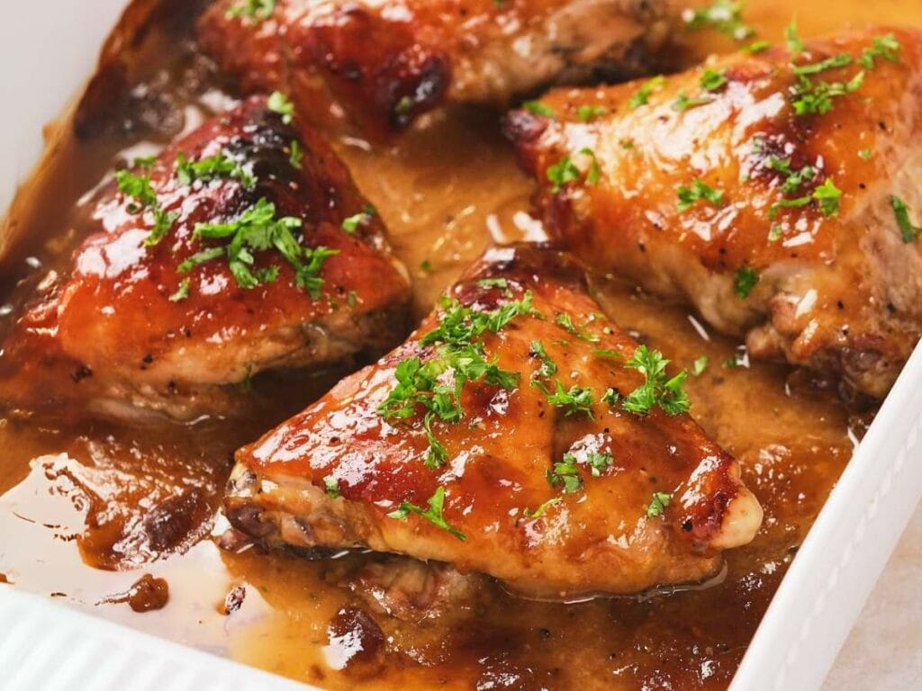 Baked apricot chicken in a brown sauce, garnished with chopped parsley, in a white dish.