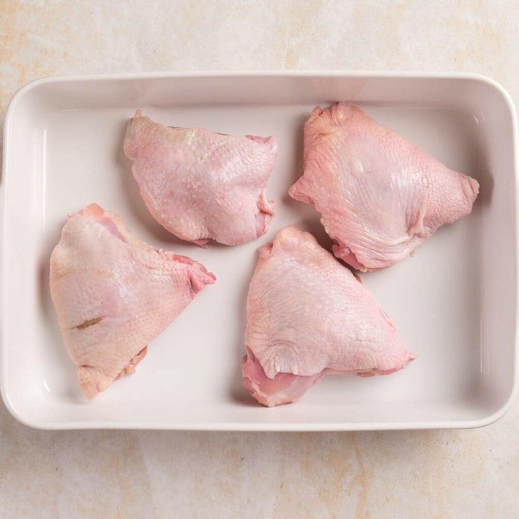 Four raw chicken thighs arranged in a white rectangular dish on a light marble surface.