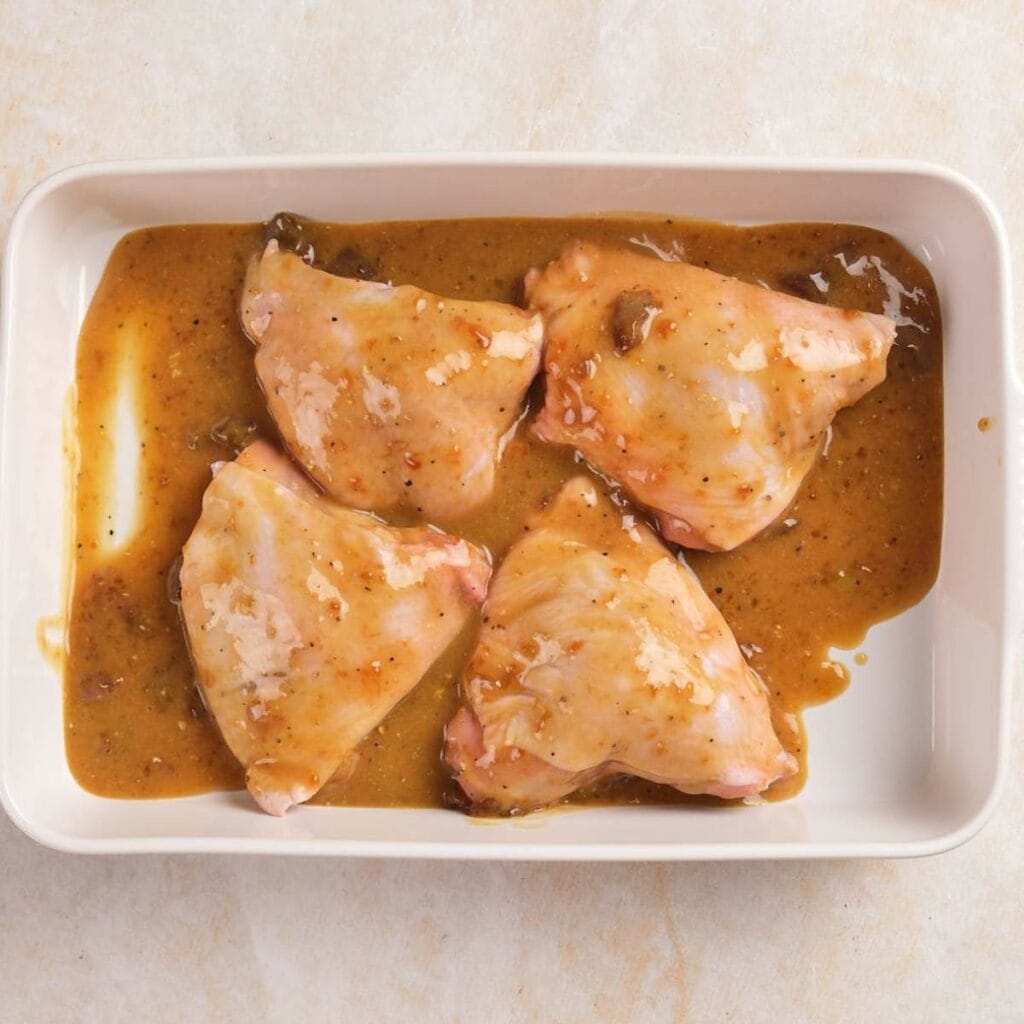 Four raw chicken thighs in a rectangular dish, marinated with a brown sauce.
