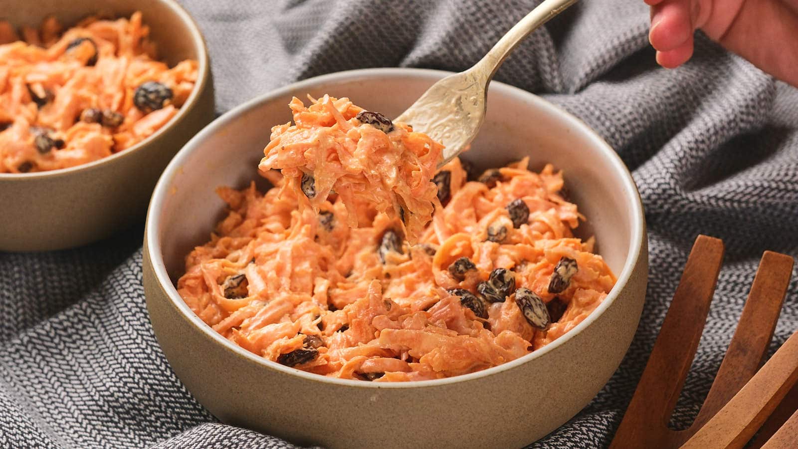 A bowl of carrot salad with raisins is on a textured gray cloth.