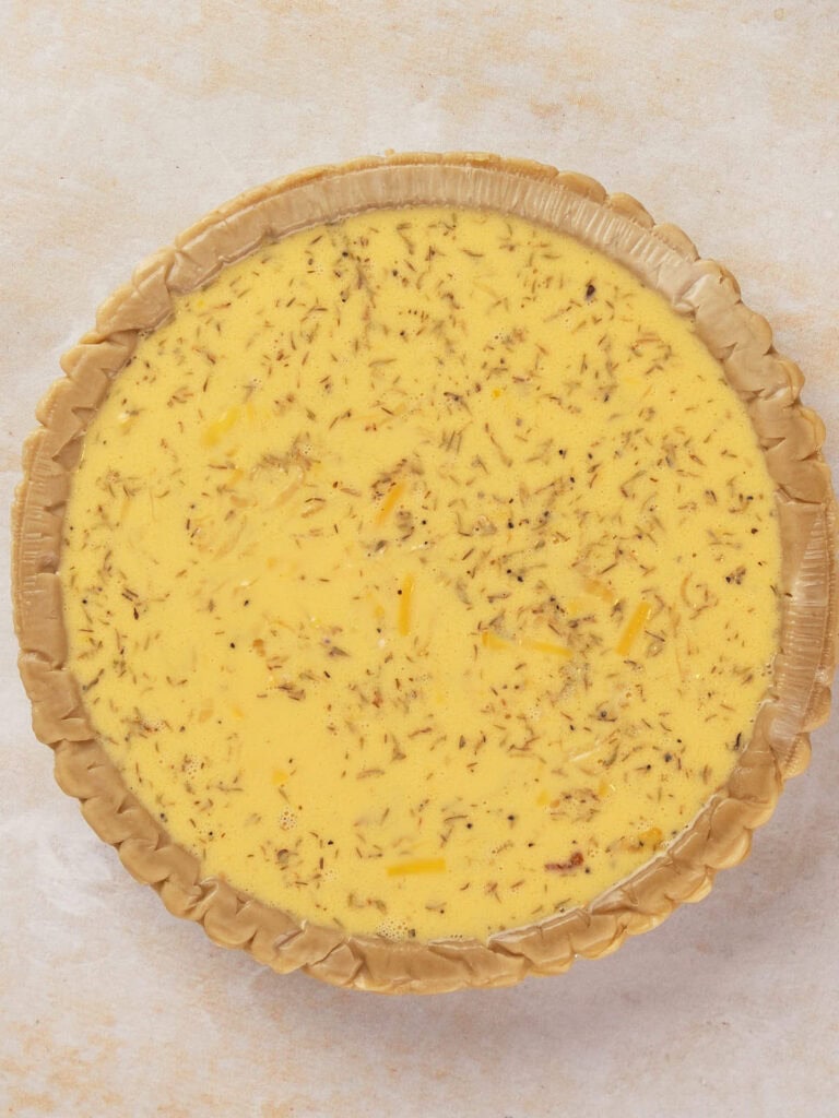 Unbaked quiche with a creamy egg mixture and herbs in a crimped pastry shell on a light-textured surface.